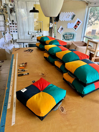 A view of the Peace Cabin studio, with a big work table stacked with colorful cushions in progress of being put finished