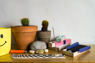 A wooden shelf with 2 small cactus, smooth river rocks, and a stack of different types of incense boxes. One stick is in a checkerboard burner, wafting incense.