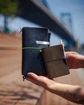 Someone off camera holds a full size and passport size TRAVELER'S notebook in both hands, one black and one olive leather. In the background is the Ben Franklin Bridge in Philadelphia