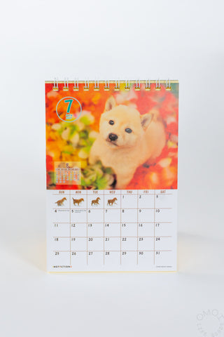 A spiral desk calendar featuring a photograph of a shiba inu in a flower bed featuring the month of July