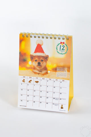 A spiral-bound desk calendar featuring a Shiba Inu dog wearing a Santa hat on the cover, with a monthly calendar below