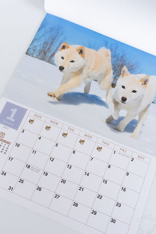 A printed calendar page showing a winter scene with two dogs running through snow under a blue sky, with a monthly calendar grid underneath 