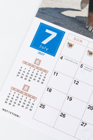 A close-up of a printed calendar page showing July 2027, with smaller weekday grids, and small dog face graphics in the calendar squares. 