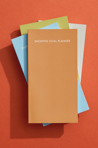 Stack of colorful planners with a focus on an orange one labeled '3 Months Goal Planner' on a red background.