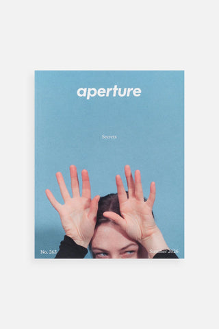 A magazine or book cover titled 'aperture' with a light blue background, featuring a person's face partially obscured by raised hands.