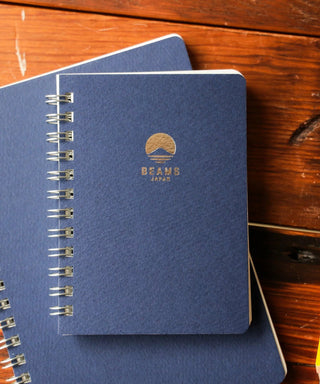 Two spiral blue notebooks with a Beams Japan logo on a wooden surface