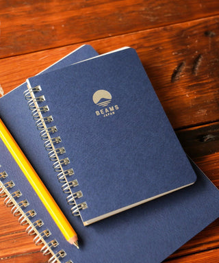 Blue notebooks with a pencil on a wooden surface, featuring the Beams Japan logo.