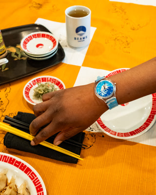 Person reaching for chopsticks around a dinner setting and wearing a blue and white watch with a mountain design