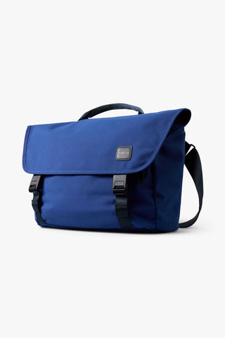 An indigo blue messenger bag with black straps and a small brand logo on the front flap, shown against a plain white background.