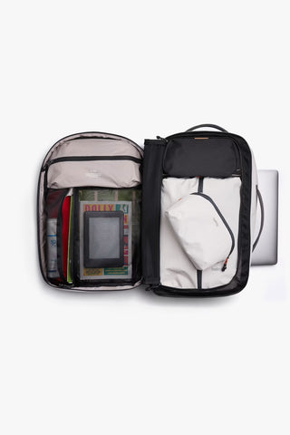 An open backpack showing multiple interior compartments filled with travel items such as a tablet, books, and a small pouch. A silver laptop is partially visible on the right side to show compartment for laptop