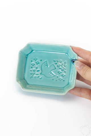 Hand holding a teal ceramic dish with decorative patterns on a white background