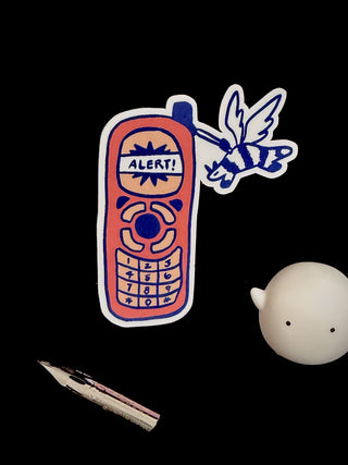 Sticker of a retro phone with 'ALERT!' text, a fountain pen nib, and a small round object on a black background.