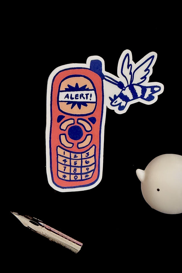 Sticker of a retro phone with 'ALERT!' text, a fountain pen nib, and a small round object on a black background.