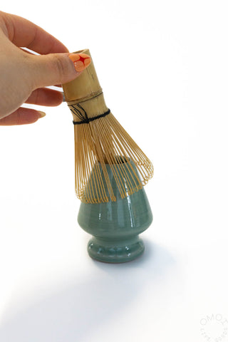 Teal ceramic whisk stand with bamboo whisk held by a hand on a white background