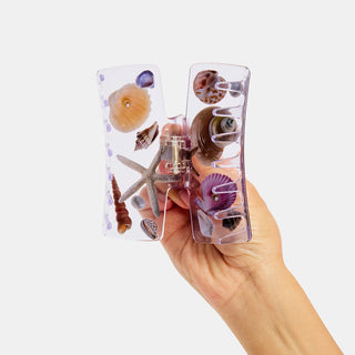 Hand holding a clear claw clip with seashell designs on a white background