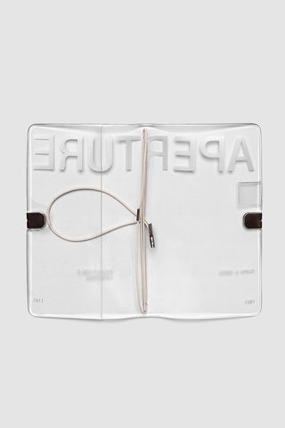 A clear acrylic notebook cover featuring embossed branding that reads 'APERTURE' and elastic closure, shown against a light gray background.