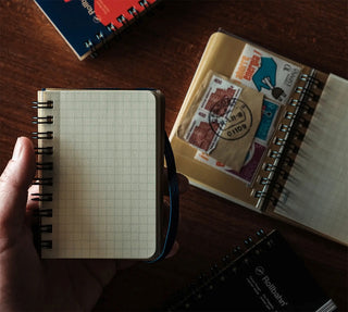 person holding pocket sized notebook with other notebook in the background that has stickers and stamps on it