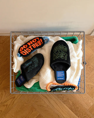 Pair of black slippers and sleep mask with text in a wire basket on a wooden floor.