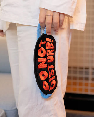Person holding a black and orange 'Do Not Disturb' sleep mask with a blue handle.