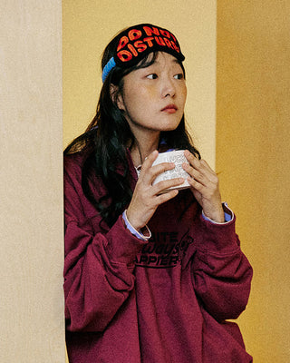 Person wearing a maroon sweatshirt and 'Do Not Disturb' sleep mask, holding a mug.