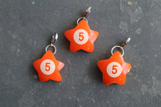 Three bright orange star-shaped charm pendants with a white circle containing the number 5, attached to silver clasps, displayed on a dark gray textured surface.