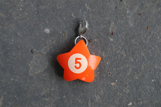 An orange star-shaped charm with a white circle in the center containing the number 5, attached to a silver clasp, resting on a dark gray textured surface.