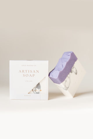 A bar of lavender artisan soap next to its white packaging box, featuring a open cut out part of the box that displays the soap inside. The box is printed with 'GOLD WATER CO.'