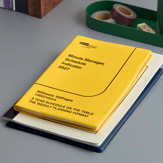A yellow 2027 Minute Manager planner notebook with black text, stacked on top of notebooks on a desk with other stationery items in the background