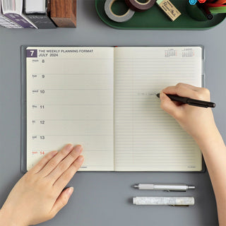 An open weekly planner with a person writing, shown on a gray desk with stationery items around it.