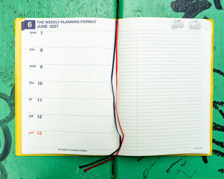 An open weekly planner with dates and lined pages, red and navy ribbon bookmarks, laid flat on a bright green textured surface.