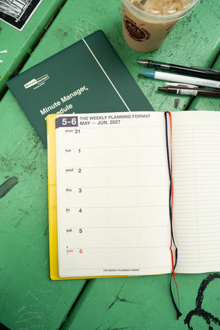 An open weekly planner with lined pages and date columns on green wooden table, next to a closed green Minute Manager planner, pens, and an iced coffee cup.