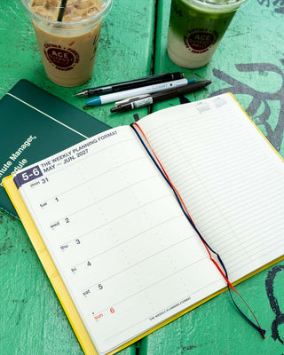 An open weekly planner with a lined page and red/blue ribbon bookmarks, styled on a green wooden surface with pens, two iced coffee drinks, next to a dark green notebook.