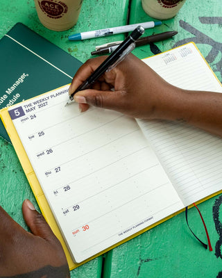 A person's hands writing in a weekly planner notebook with a black mechanical pencil, on a bright green wooden table alongside coffee cups and pens.