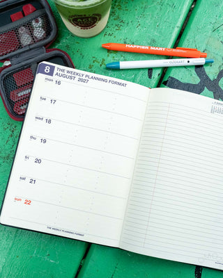 An open weekly planner showing August 2027 in a weekly layout format on a green wooden surface with pens, a cup, and a small stationery kit
