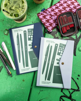 Two planner covers displayed on a wooden green tabletop, alongside pens, a pink patterned pouch, a red stationery kit set, and an iced matcha drink.