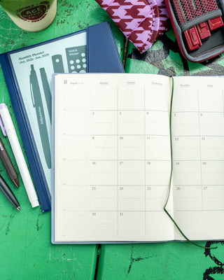 An open monthly calendar planner on a green wooden surface, surrounded by stationery items including pens, a patterned pouch, and a red stationery kit