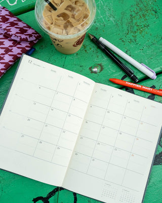 An open monthly planner on a wooden green tabletop, surrounded by iced coffee, pens, and a patterned fabric case.