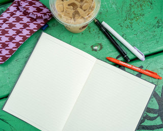 An open planner with lined pages on a green wooden surface, surrounded by a pink-and-brown patterned pencil case, an iced coffee cup, and pens.