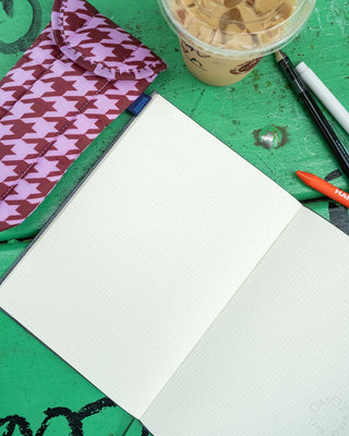 An opened planner with grid page on a wooden green table, surrounded by a houndstooth case, pens and pencil, and an iced coffee cup.