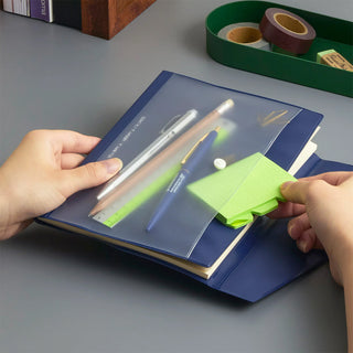 A navy blue pocket notebook with a clear plastic front cover being held open by two hands. A bright green sticky note is tucked into the pocket, with pens and other stationery visible inside.