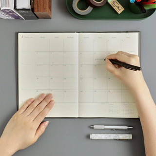 Top-down view of a person writing in an open monthly planner with a black pen on a gray desk, surrounded by stationery items.