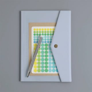A light planner with a triangular flap and a single metal snap button, shown closed on a gray background. Inside the folder is a sheet of round stickers and a silver pen across it.
