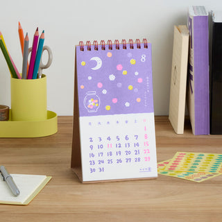 A spiral-bound desk calendar with a purple decoration with small stars and the moon. On the desk in the background is other stationery items such as a cup with colored pencils and books.