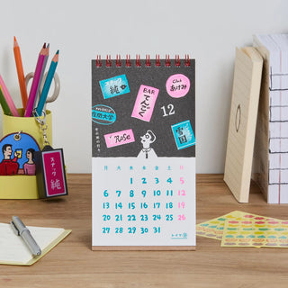 A spiral-bound desk calendar with Japanese text and colorful retro style design on a wooden desk next to pencils, sticky notes, and notebooks.