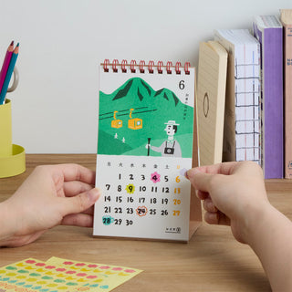 A person's hands are holding a small spiral-bound desk calendar with a colorful illustration of mountains and cable cars. The calendar is placed on a wooden desk with books and stationery in the background.