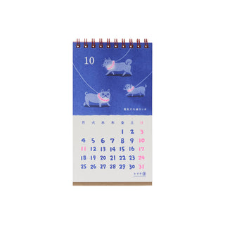 A spiral-bound desk calendar page with a blue top section showing three illustrated dogs wearing pink collars, and a white bottom section with a numbered calendar grid for October