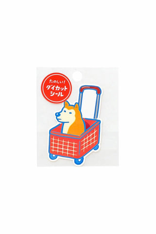 A die-cut sticker featuring an orange Shiba Inu dog sitting in a red shopping cart with blue wheels and handle
