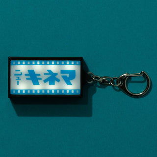 A rectangular keychain with a glowing white sign displaying blue Japanese text and filmstrip borders, attached to a silver metal clasp, set against a solid blue background.