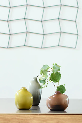 Three ceramic vases in green, gray, and brown on a surface with a geometric patterned curtain in the background.