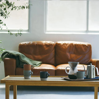 Brown leather sofa with a wooden coffee table featuring mugs and a coffee maker in a bright room.